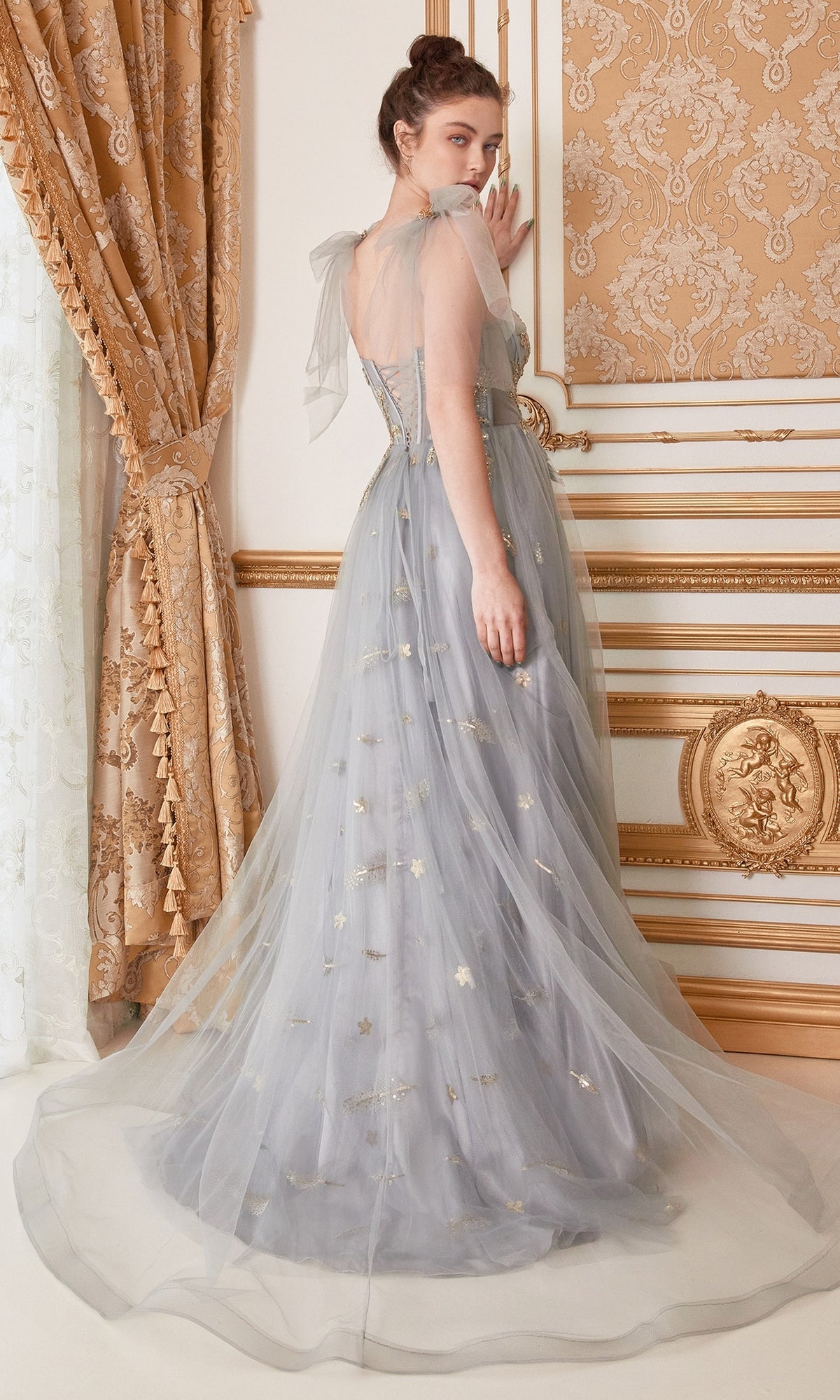 A woman in the Shoulder-Bow Long Corset Prom Ball Gown A0824—a sheer, light blue gown with gold accents and corset bodice—stands in an ornate gold and cream room, looking over her shoulder.